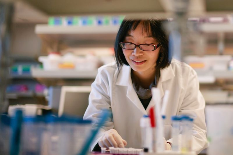 Scientist in lab coat works with equipment; lab setting with shelves of supplies.