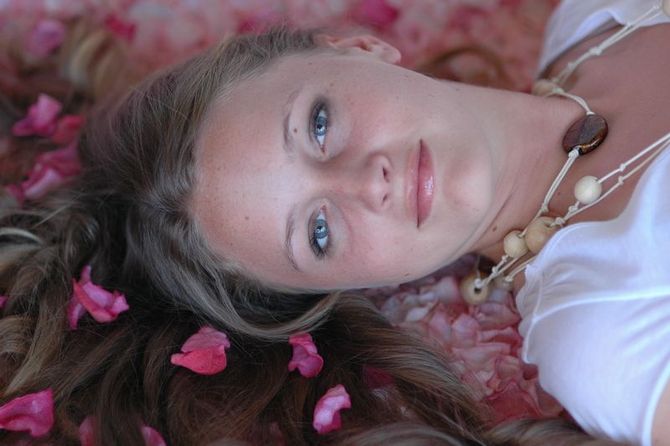 Woman with light hair lies in pink flower petals, wearing a white top and necklace; eyes gazing toward the viewer.