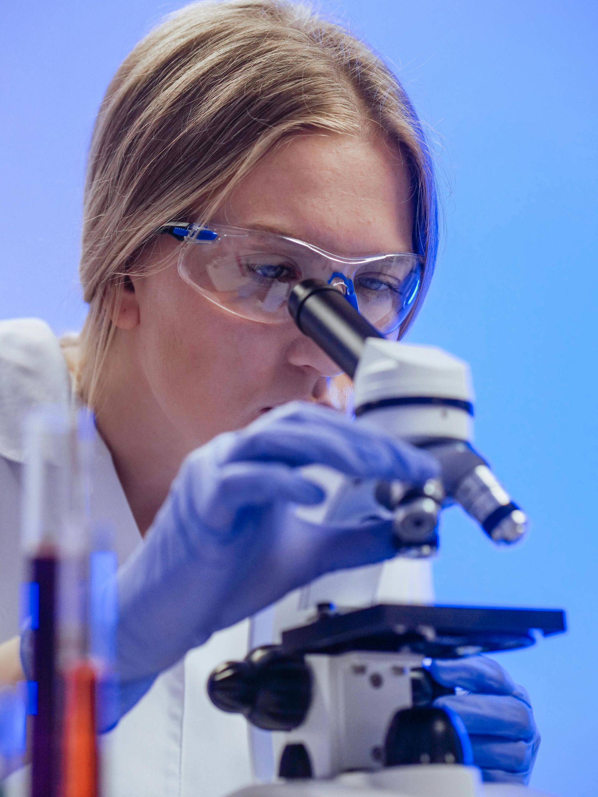 Woman in lab coat and safety glasses looking through a microscope in a laboratory setting.