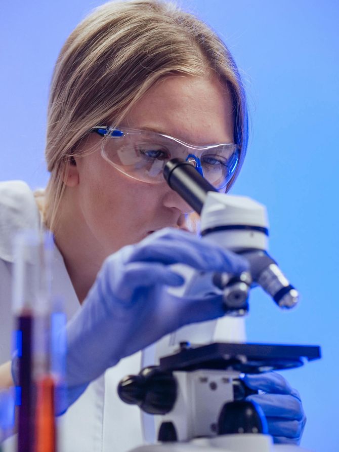 Woman in lab coat and safety glasses looking through a microscope in a laboratory setting.