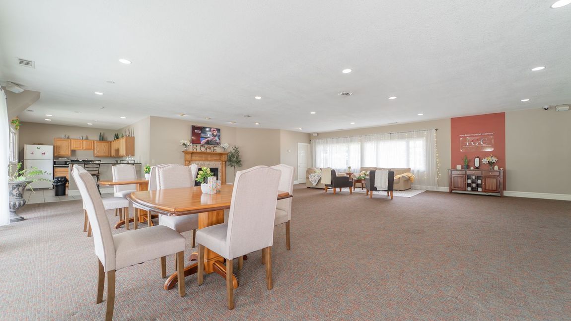 Large room with dining tables, chairs, kitchen, and seating area. Brown carpet, neutral walls, natural light.