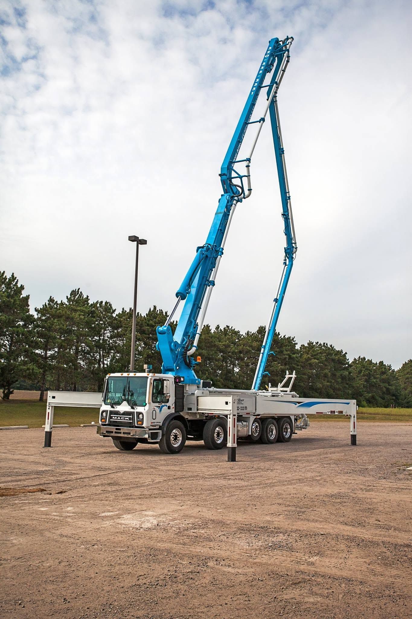 Blue boom truck with extended lifting arm in an outdoor gravel lot near trees and a light pole