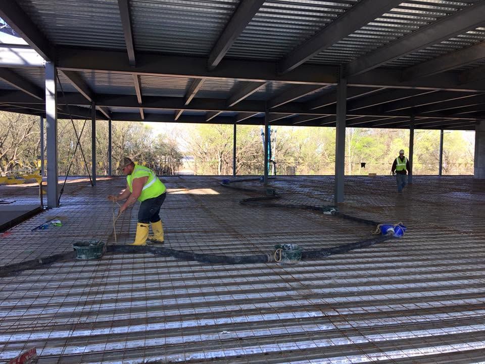Workers laying concrete in a metal-roofed construction site, one in a neon vest in the foreground