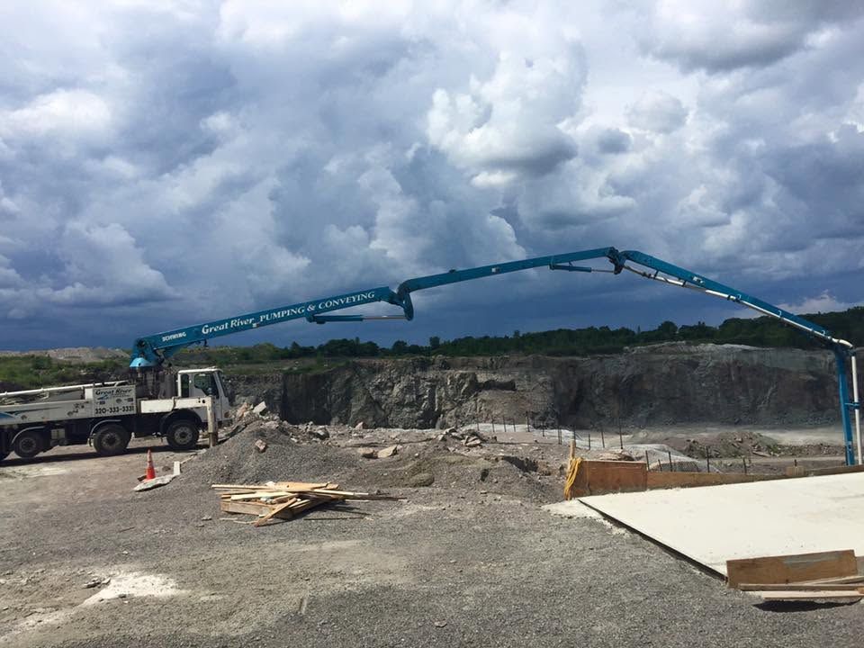 Construction site with blue concrete pump truck, gravel ground, and cloudy sky over a large excavation pit