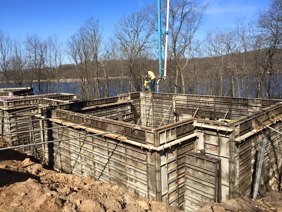 Construction site with wooden formwork and two workers on a concrete foundation near a wooded area