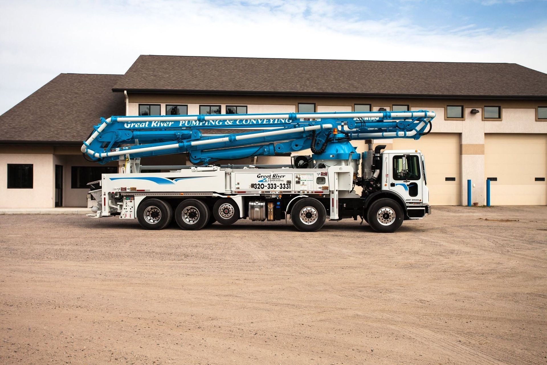 Blue concrete pump truck parked outside a beige building on a gravel lot