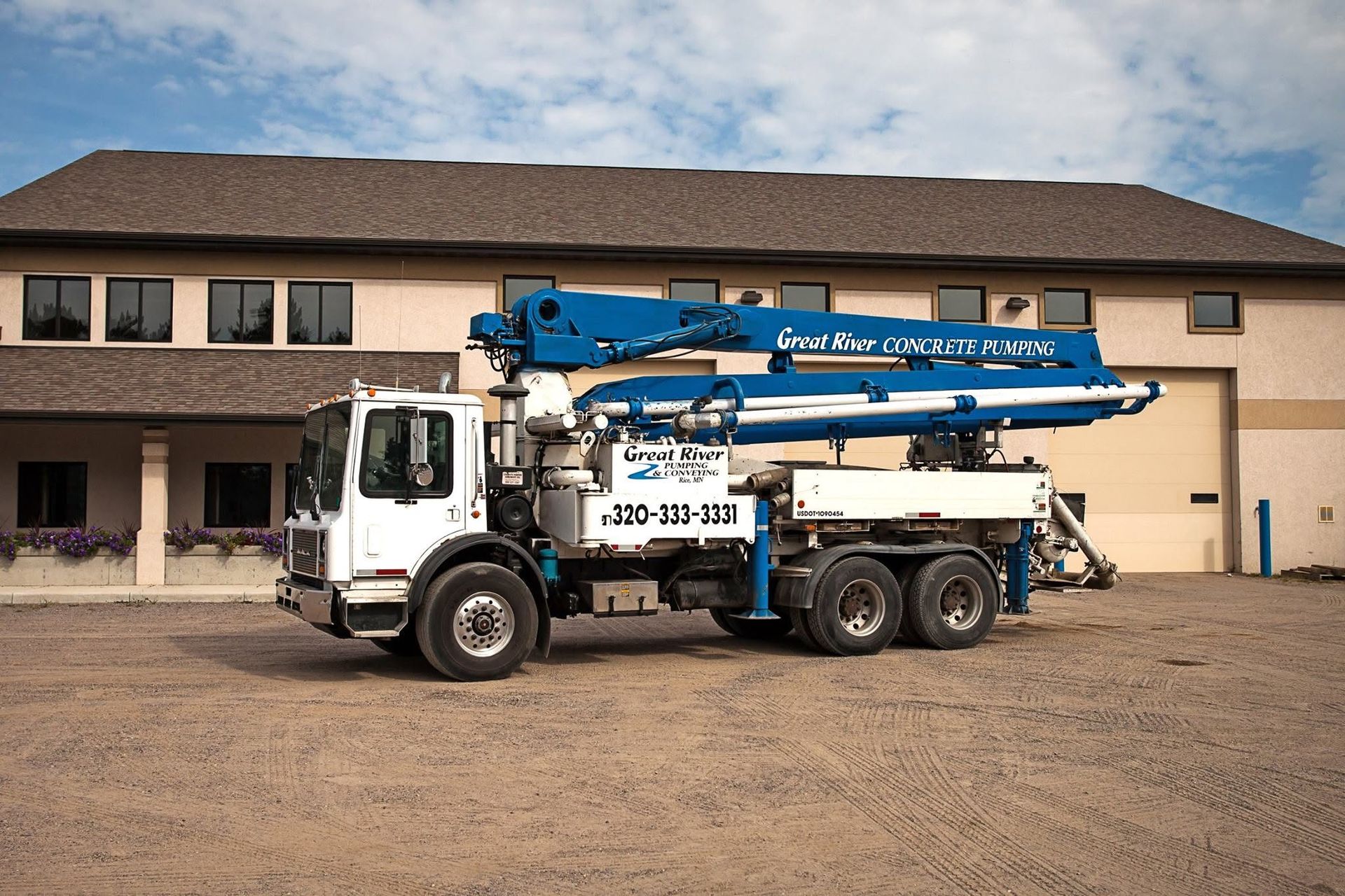 Blue concrete pump truck parked in front of a beige building on a gravel lot