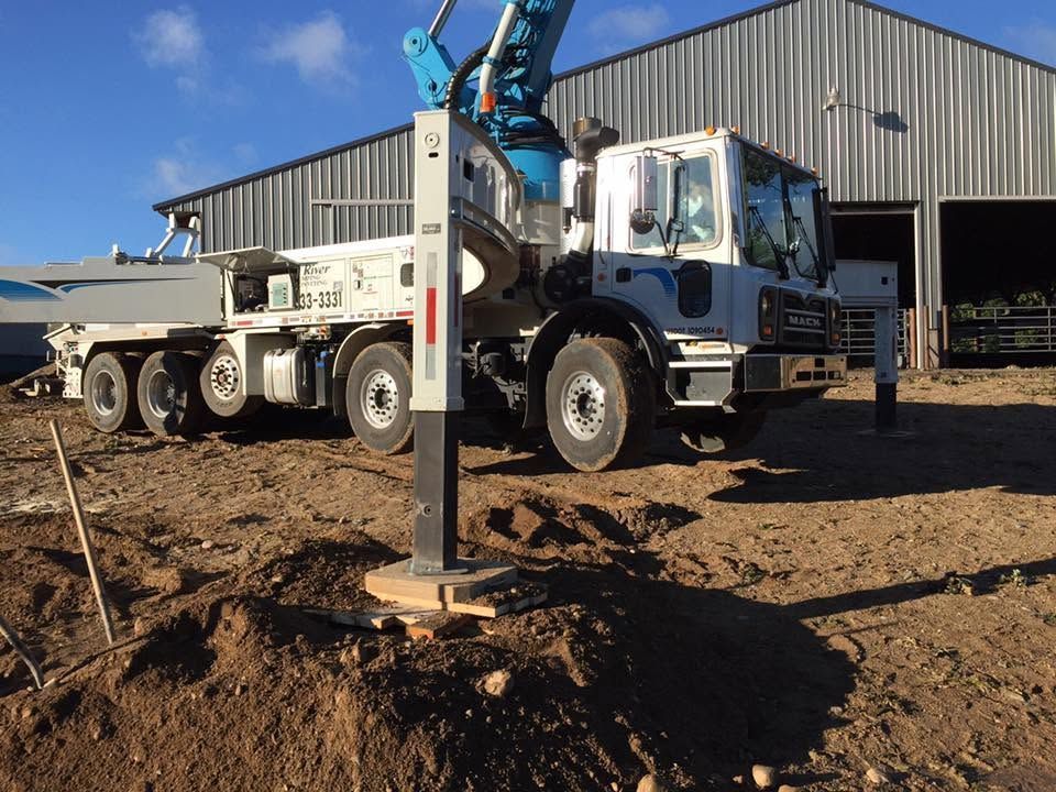 White truck with a mounted drilling rig on a dirt lot beside a metal building under blue sky