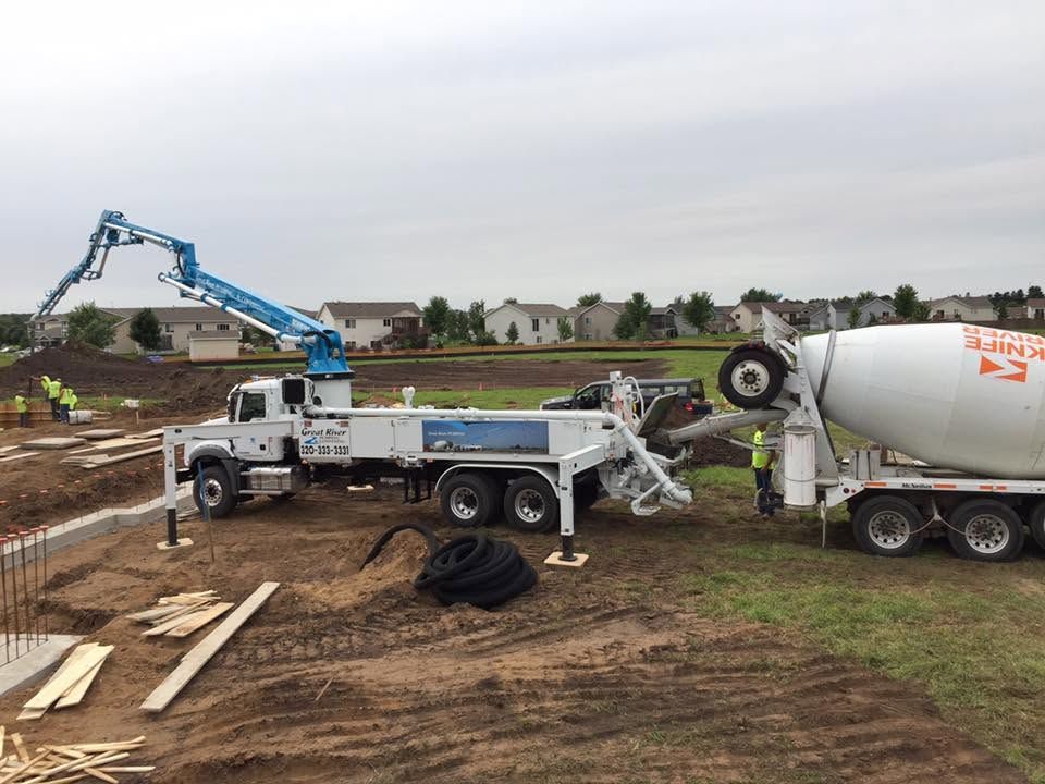 Concrete truck tipping at a construction site while a blue crane truck stabilizes it amid muddy ground and debris