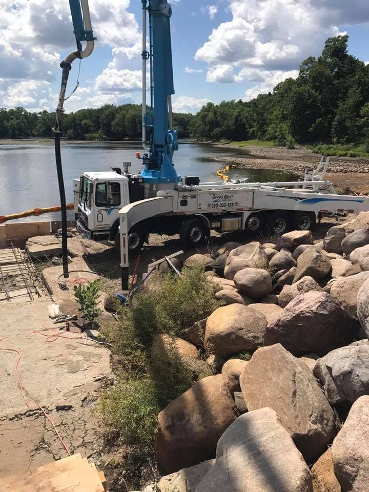 Crane truck unloading rocks at a riverbank beside a concrete dock under a blue sky.