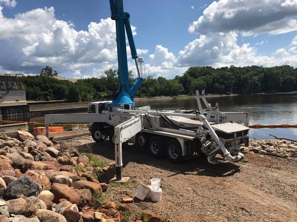 Damaged white trailer crane by a river, with a blue boom arm and debris on rocky ground