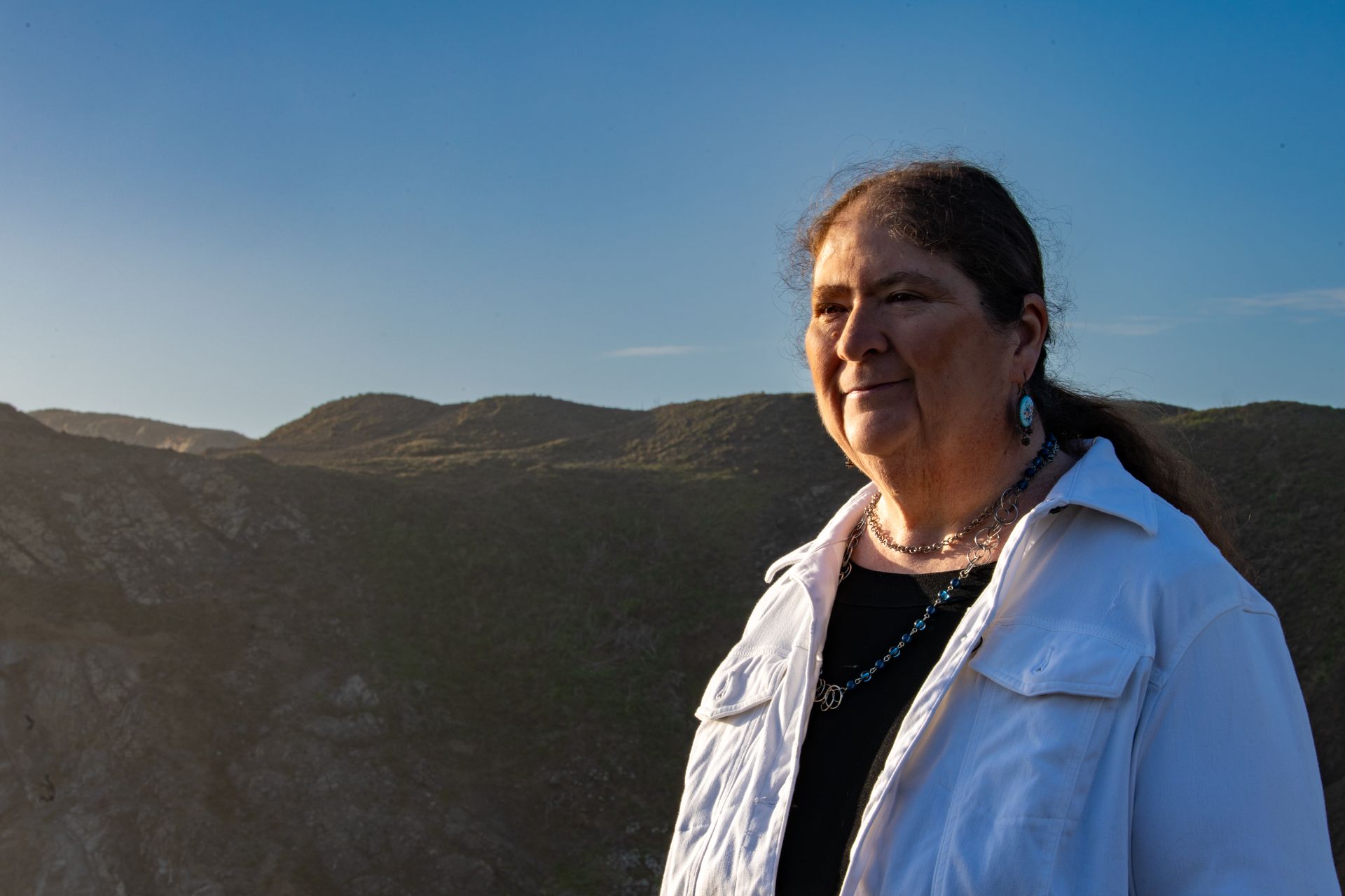 Woman in a white jacket stands outdoors with mountains under a clear blue sky.