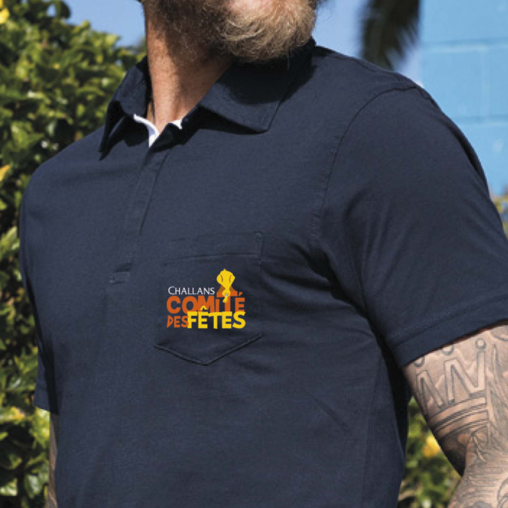 Homme portant un polo bleu marine avec un texte et d'un logo orange et jaune. Il a une barbe et un tatouage sur le bras.