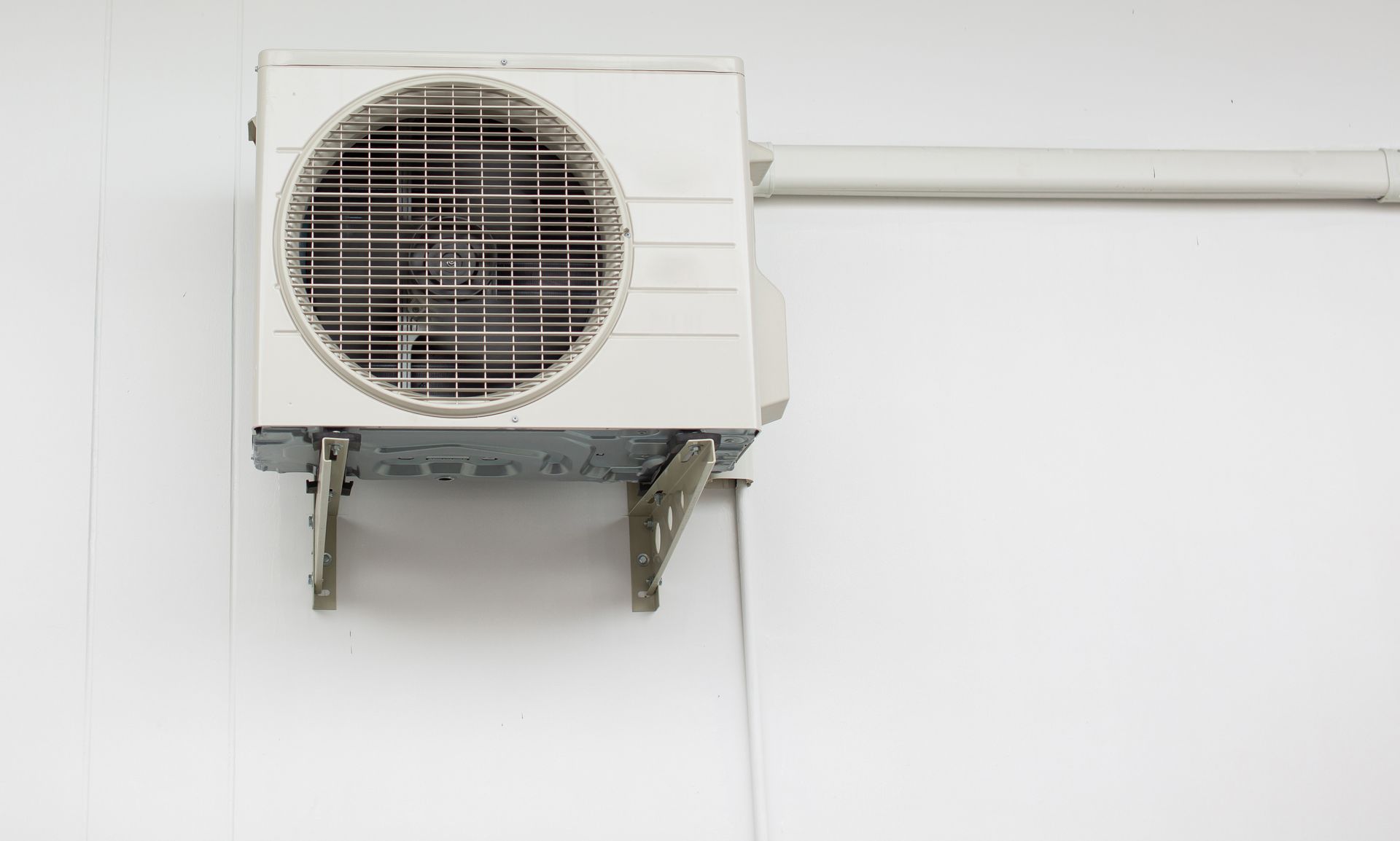 Wall-mounted outdoor air conditioning unit with visible piping on a plain white wall.