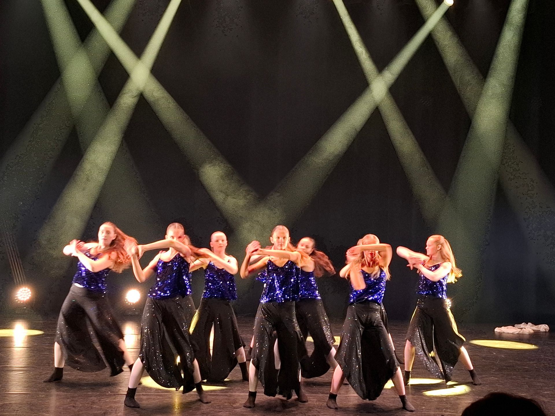 Dansere i blå paljetttopper og svarte bukser opptrer på scenen under sterke spotlights.