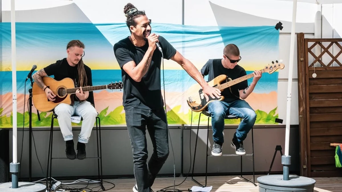 Band opptrer utendørs; sanger med dreadlocks, gitarist og bassist spiller mot et strandlandskap i bakgrunnen.