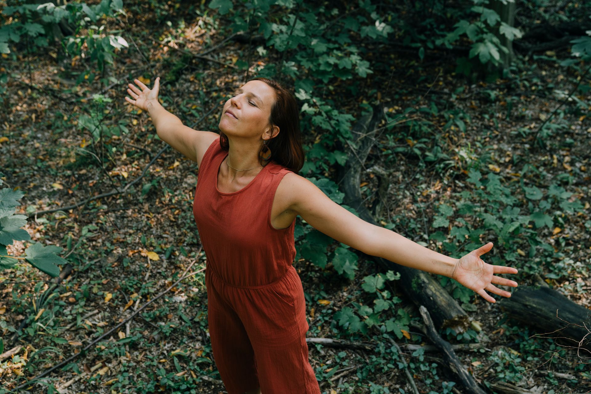 Hannah Bunk in einem roten Overall mit ausgestreckten Armen und geschlossenen Augen in einem Waldgebiet.
