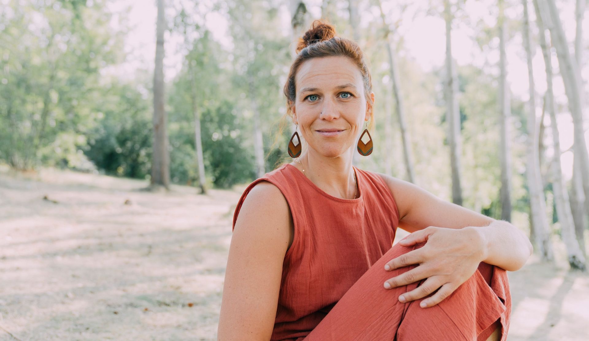 Hannah Bunk, im orangefarbenem Outfit, sitzt lächelnd im Freien, im Hintergrund sind Bäume zu sehen.