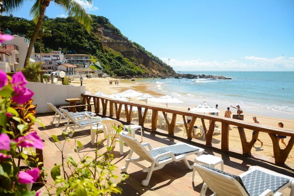 Espreguiçadeiras e corrimão de madeira à beira-mar com vista para uma baía ensolarada e flores cor-de-rosa em primeiro plano.