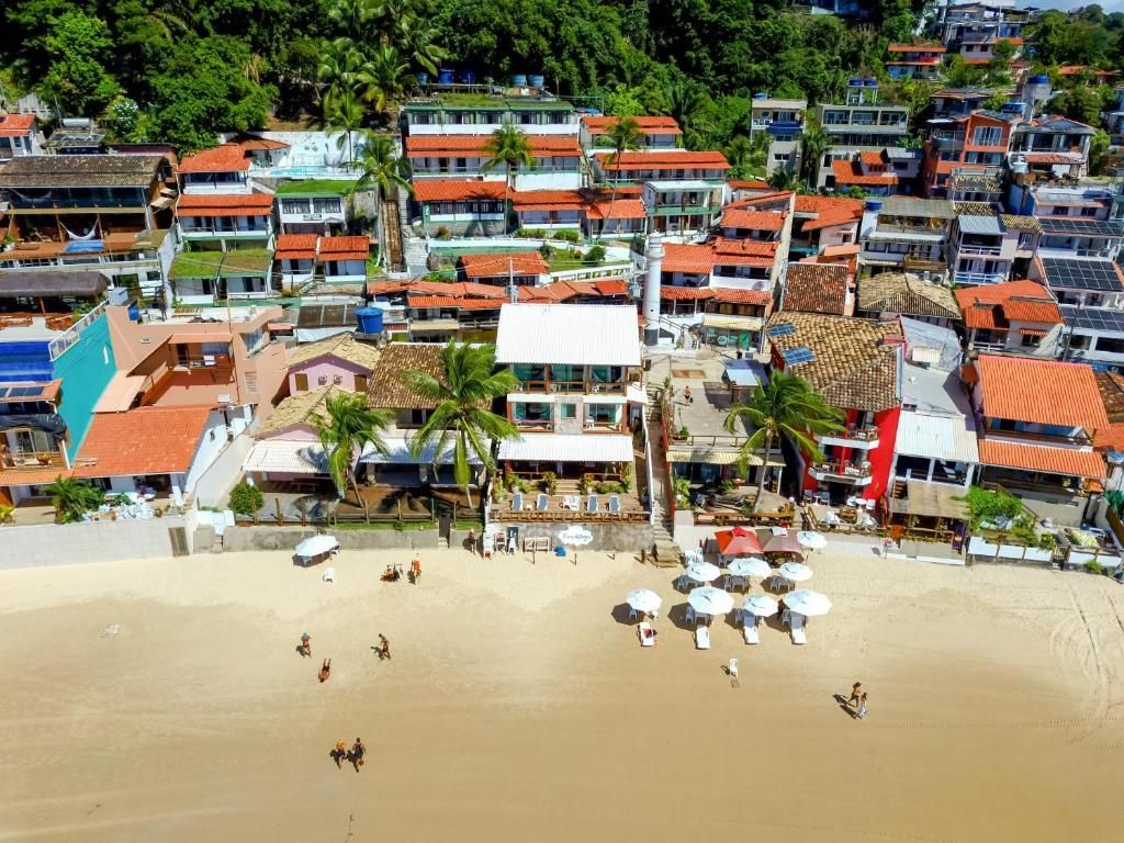 Vista aérea de uma praia tropical com guarda-sóis, banhistas e construções coloridas à beira-mar, tendo as árvores como pano de fundo.