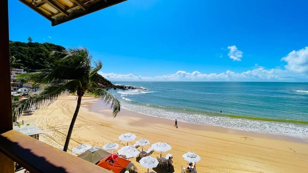 Praia tropical ensolarada com areia dourada, ondas turquesa, guarda-sóis brancos e uma palmeira sob um céu azul.