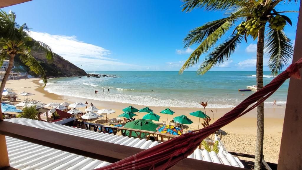 Praia tropical ensolarada com água turquesa, palmeiras, guarda-sóis e pessoas na orla.