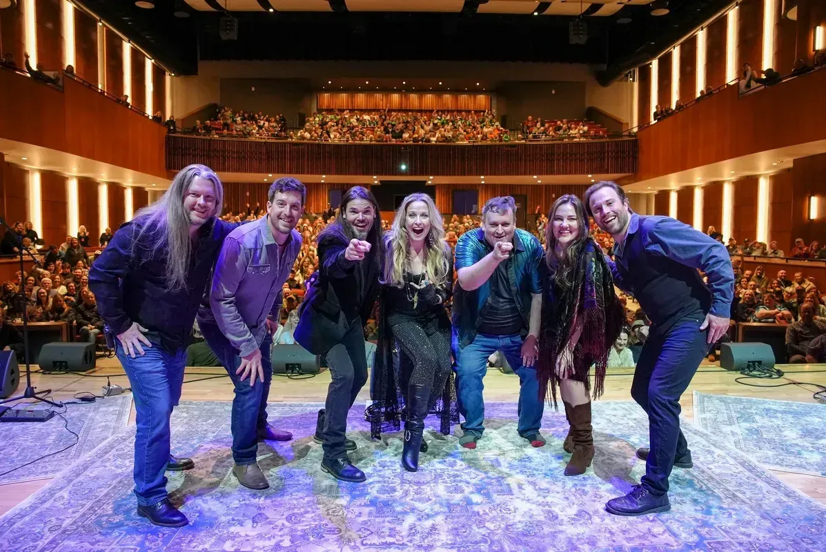 Six performers on a theater stage, leaning toward the camera under blue and purple lights.