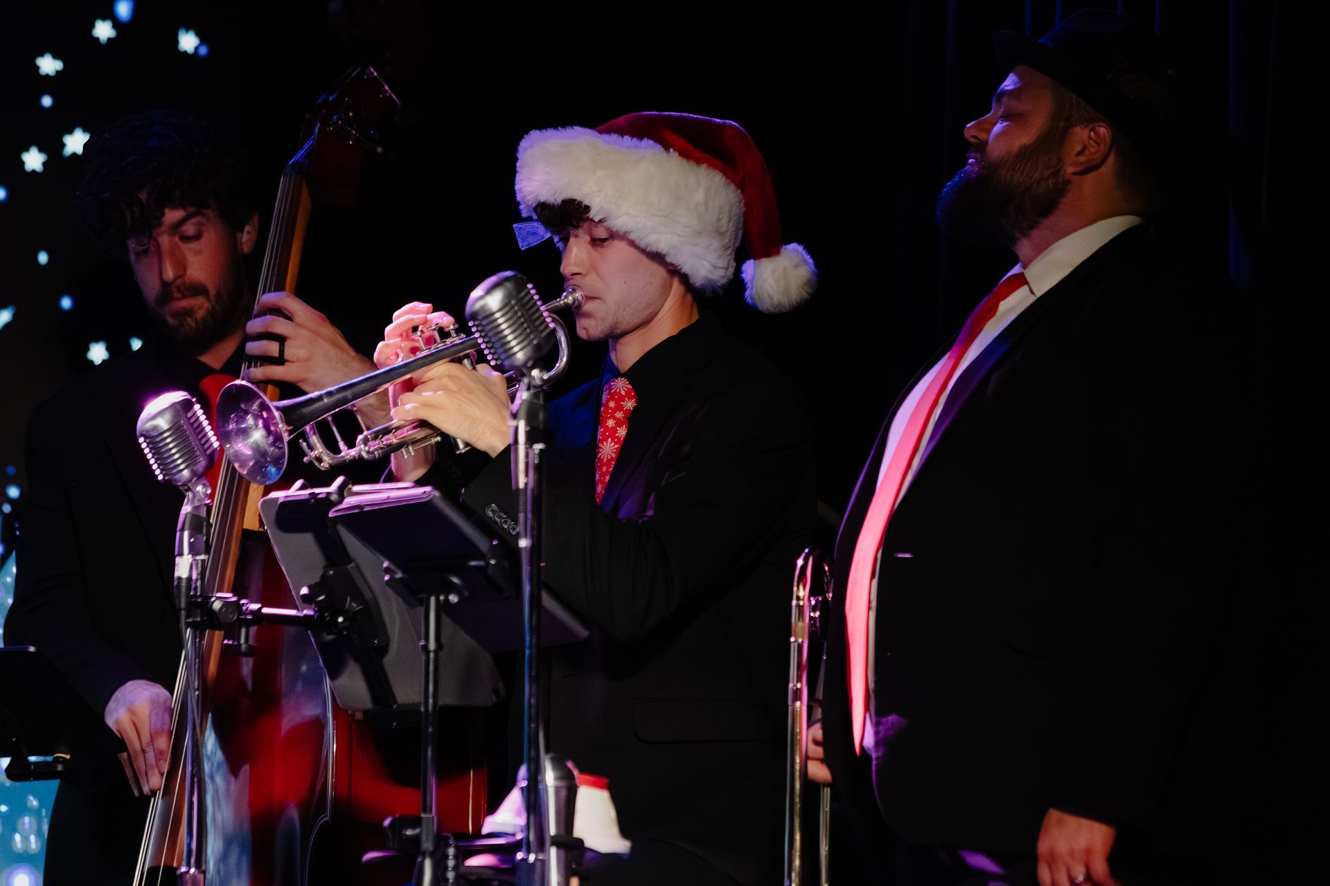 Musicians performing on stage, one wearing a Santa hat and playing a trumpet beside microphones