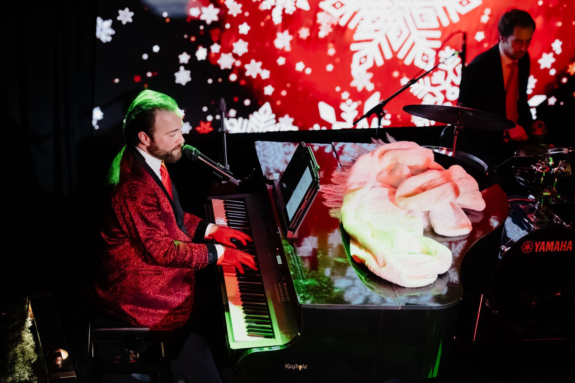 Singer in red jacket playing piano on stage with red snowflake backdrop and a large white flower display