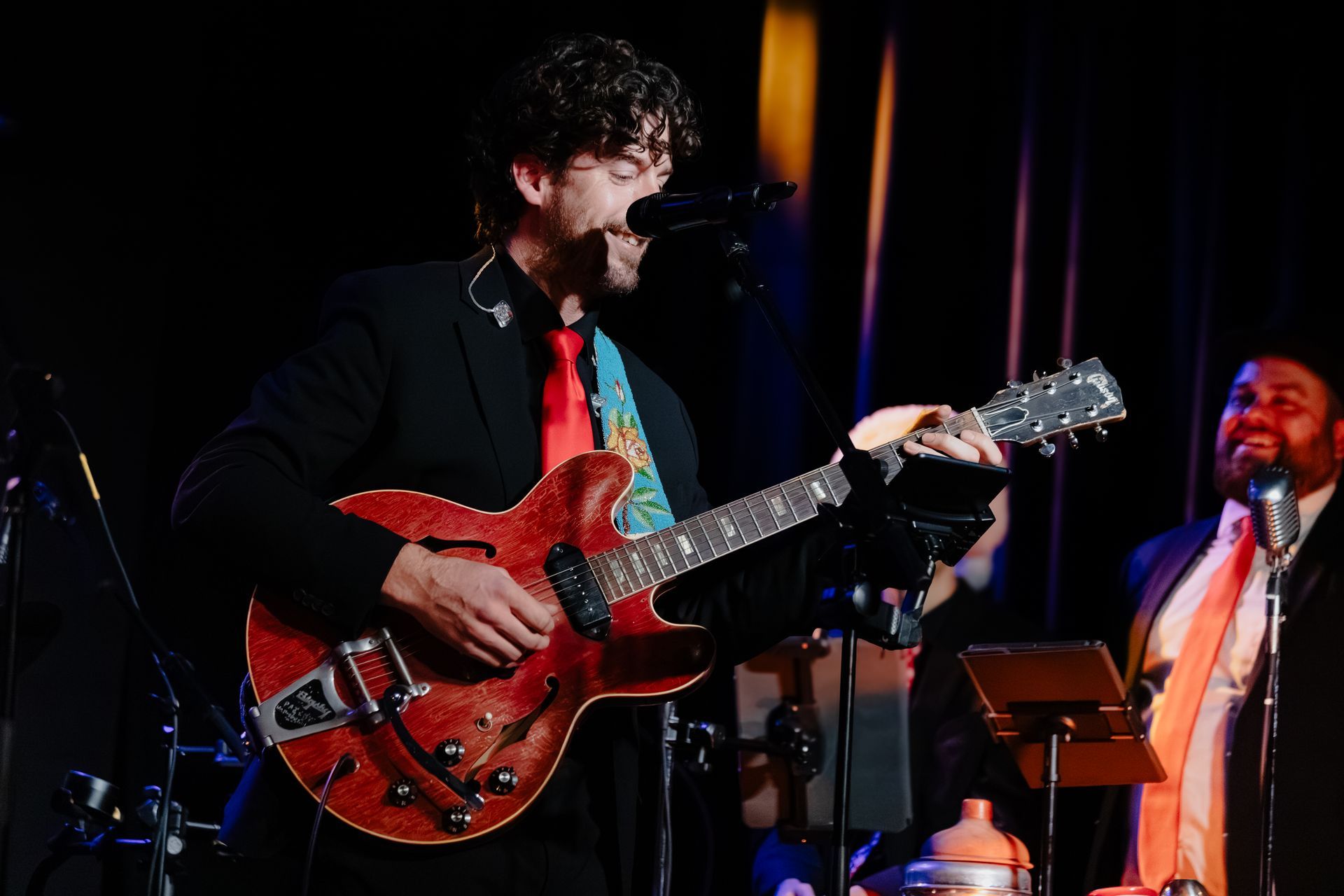 Musician singing into a mic while playing a red guitar on a dimly lit stage