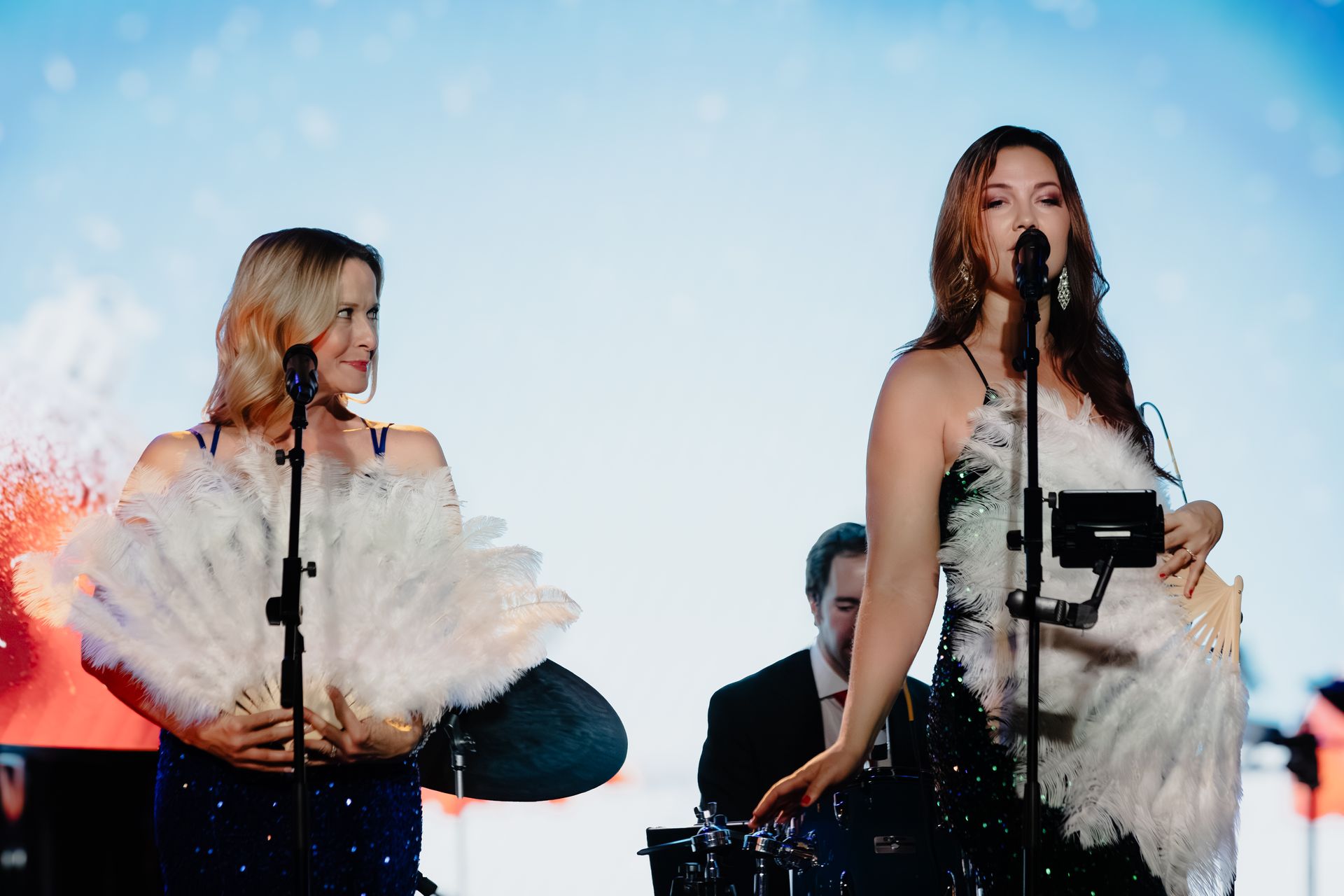 Two singers perform onstage with microphones, wearing white feathered outfits against a bright blue background.