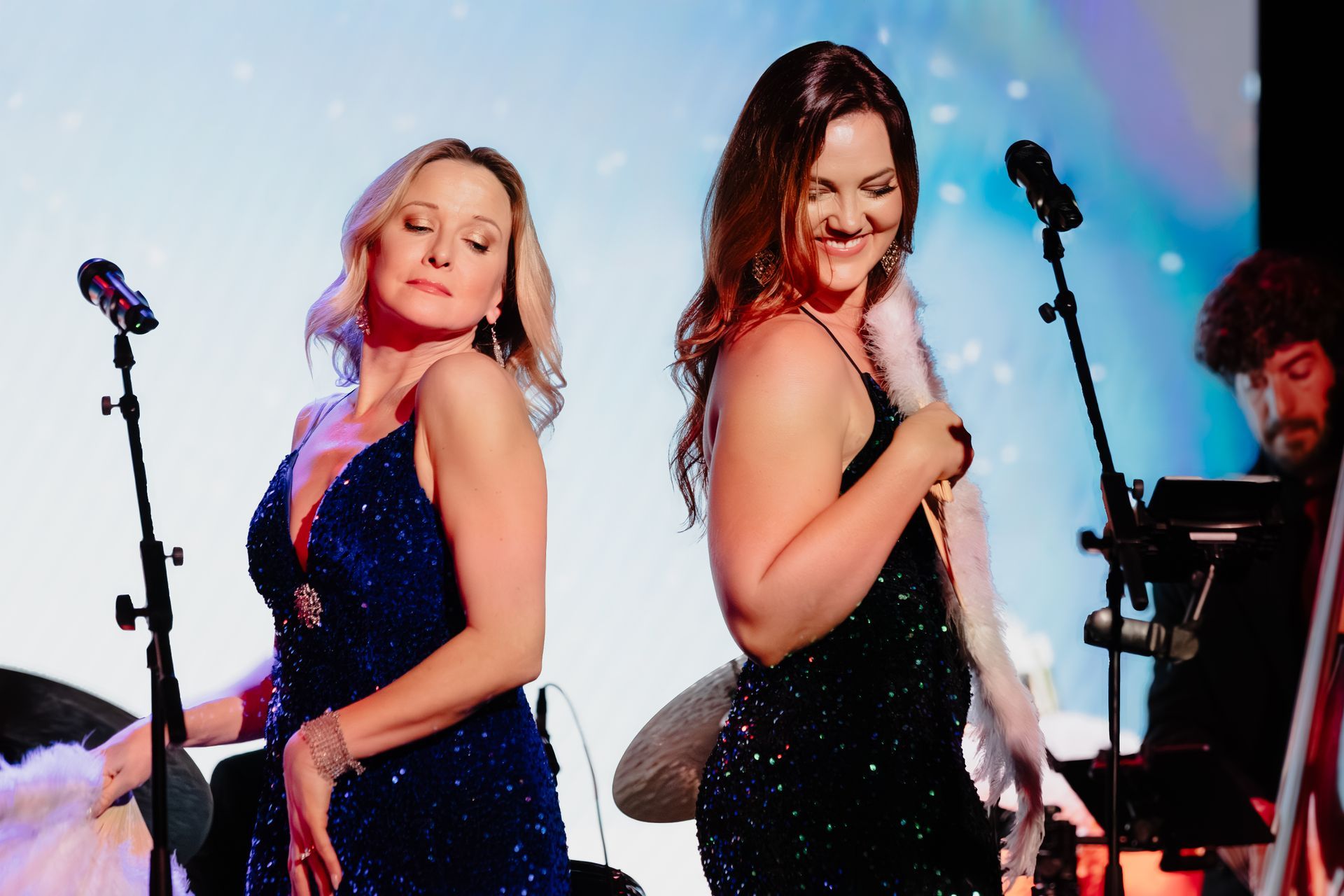 Two singers in sparkly dresses performing on stage with microphones and a drummer in the background