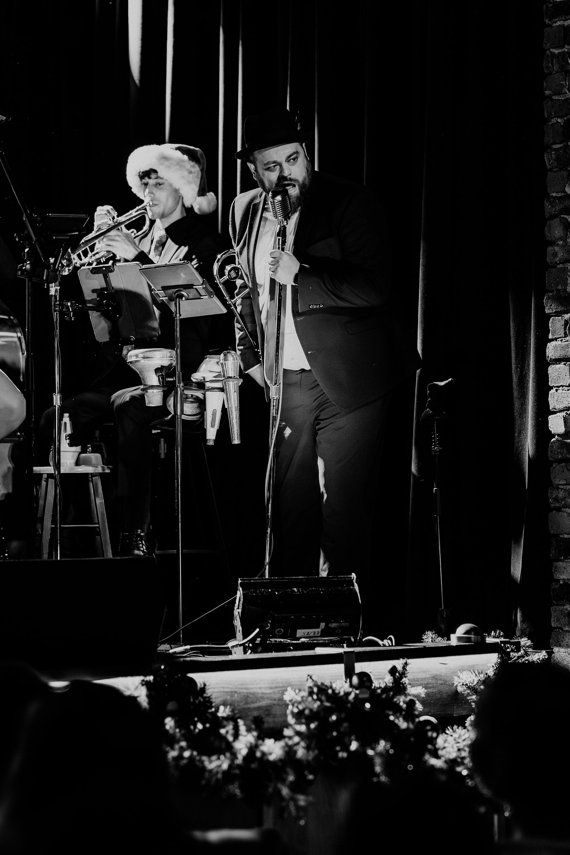 Two musicians performing on a dimly lit stage, one at a microphone and one seated at drums.