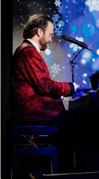 Man in red sweater playing piano and singing into a microphone on a blue-lit stage