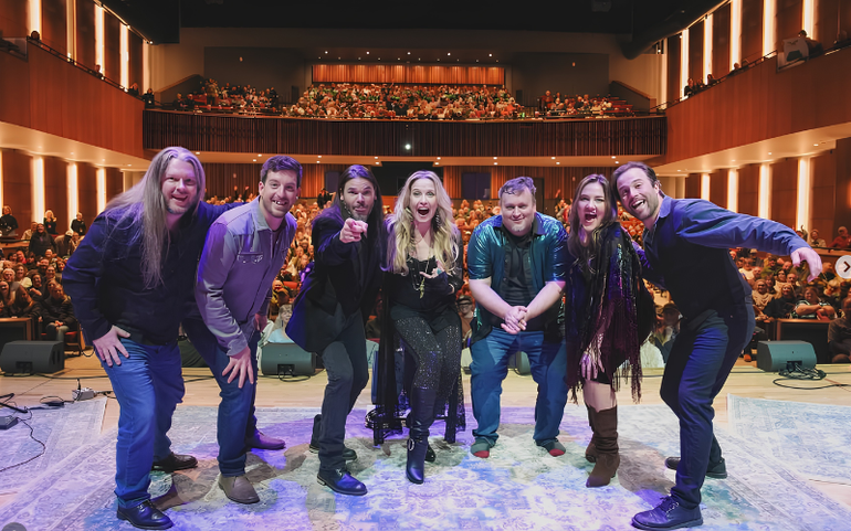 Six musicians pose onstage in a concert hall, smiling under warm lights with seats full behind them.
