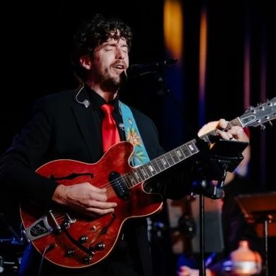 Musician singing and playing a red electric guitar onstage under warm lights