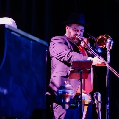 Trombone player in a black hat performing onstage under purple lighting