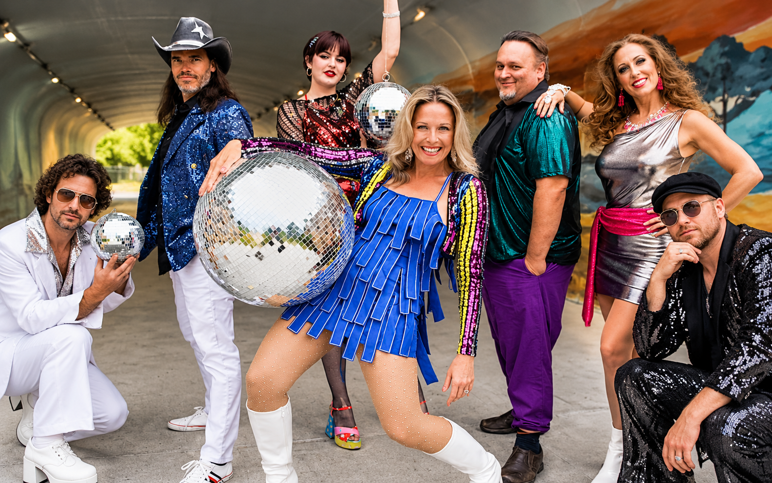 Group posing in colorful 1970s-style disco outfits, holding mirrored balls in a festive tent