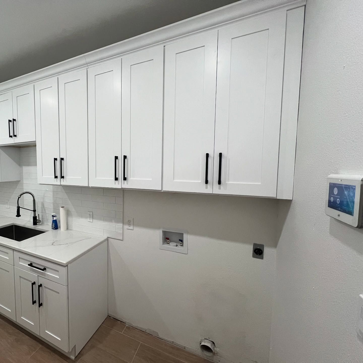White cabinets and countertop with a sink in a laundry room, featuring plumbing hookups and a smart home wall panel.