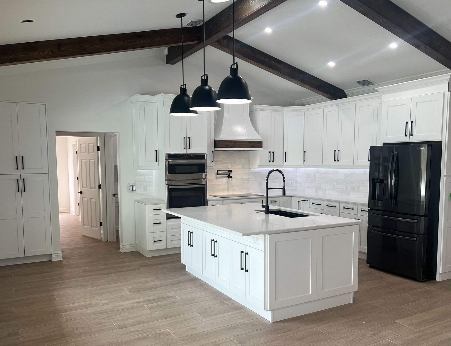 Modern white kitchen with a center island, dark pendant lights, black refrigerator, and exposed wooden ceiling beams.