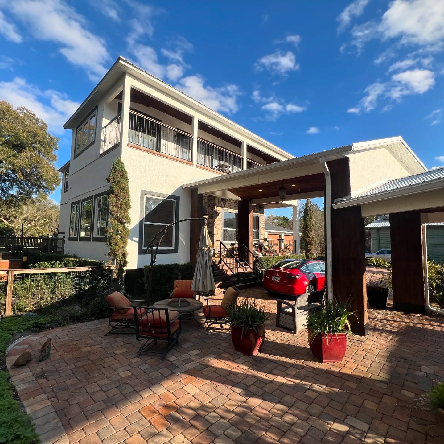 Two-story house with red car in carport and brick patio with chairs under a blue sky.