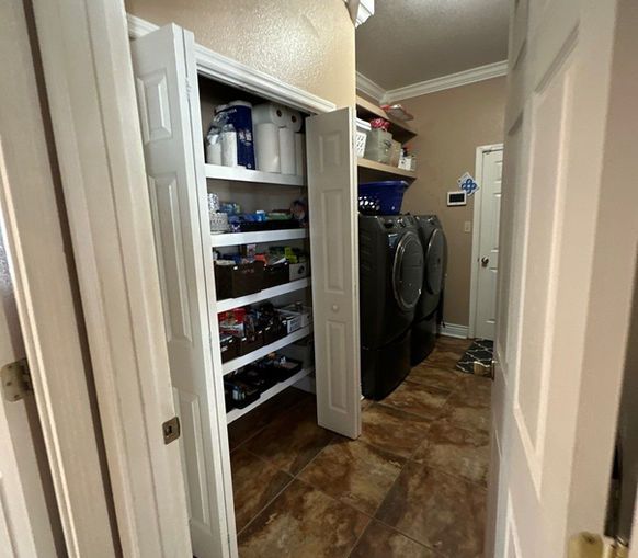 A laundry room with a white shelving unit built into a wall niche and two dark grey appliances next to a white door.