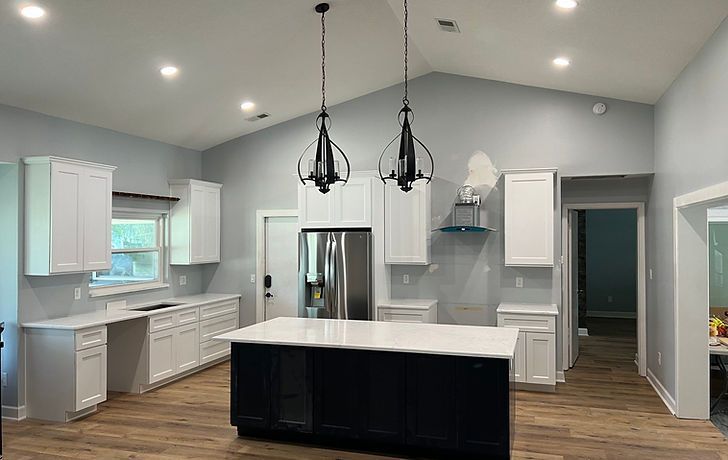 Modern kitchen with white cabinetry, a black island, two pendant lights, and light wood flooring under a vaulted ceiling.