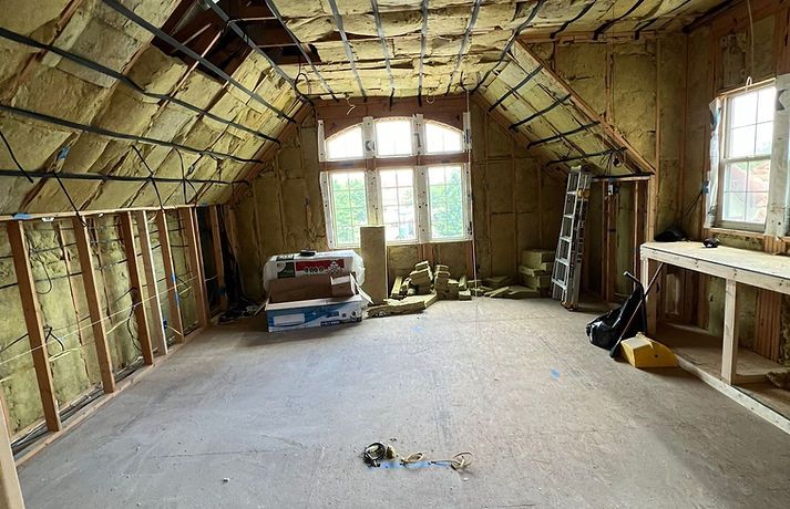An unfinished attic room featuring exposed wooden studs, yellow insulation, a central arched window, and a workbench.