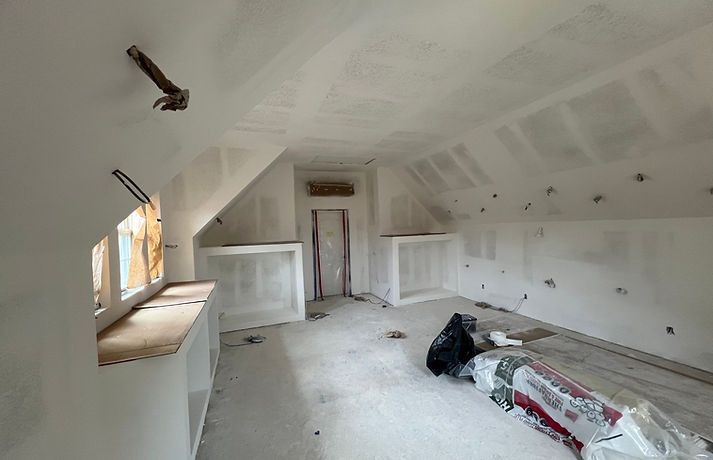 An unfinished attic room with newly installed drywall, angled ceilings, built-in shelving, and construction debris.