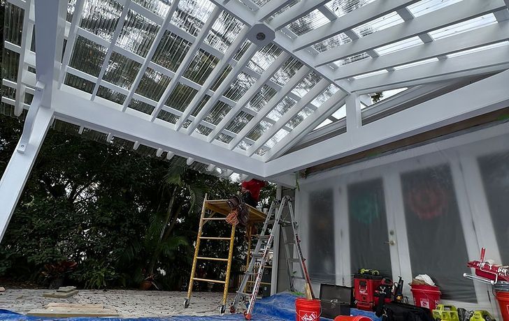 A person on scaffolding works on the ceiling of a white wooden pergola attached to a building, with tools on the ground.