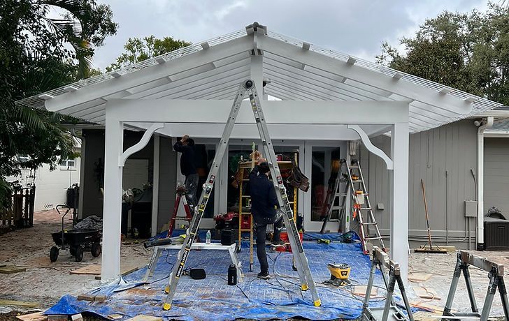 Construction workers stand on ladders installing a white wood patio cover over a blue tarp-covered yard.