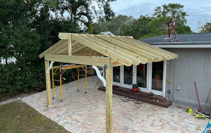 A wooden pergola frame under construction on a paved patio attached to the side of a house.