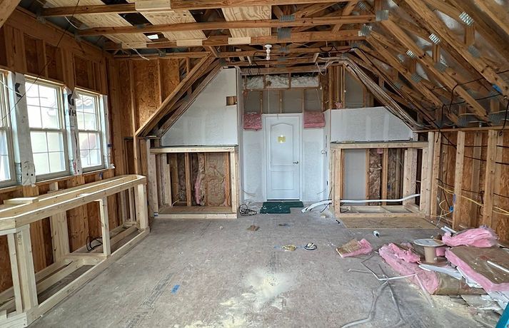 An unfinished attic room featuring exposed wooden framing, bare plywood walls, a white door, and pink fiberglass insulation.