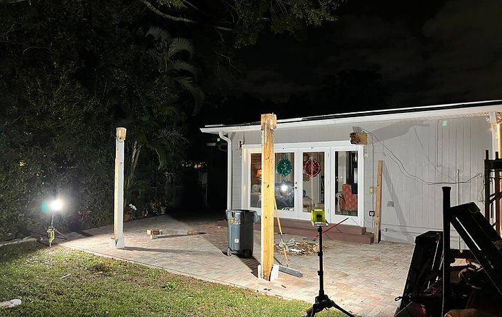 Two vertical wooden posts stand on a paved patio in front of a house at night, illuminated by portable work lights.
